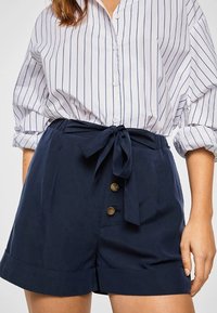 Náměsové modré šortky s vysokým pasem z hladkého materiálu, s uvázaným pasem, dvěma předními knoflíky a ohrnutým lemem. Nad nimi pruhovaná bílá košile.