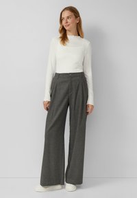 Graue weite Hose aus strukturiertem Stoff, mit einem leichten Faltenwurf an der Vorderseite und einem dunklen Knopf am Taillenbund. Kombiniert mit einem enganliegenden weißen Langarmoberteil.