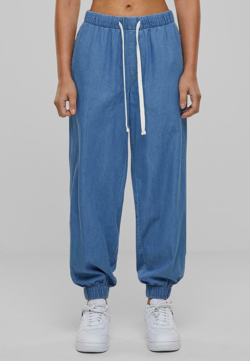 Pantalones jogger de denim, azul claro, con cinturilla elástica y cordón blanco. Con puños en los tobillos, combinados con zapatillas blancas. Diseño sencillo.