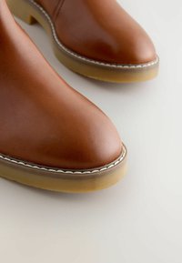 Close-up van bruin leren enkellaarsjes met lichtbruine rubberen zolen en witte stiksels rond de randen op een lichte achtergrond.