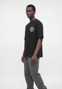 Mann trägt schwarzes T-Shirt mit Grafik und graue Cargohose, steht vor einem schlichten weißen Hintergrund.