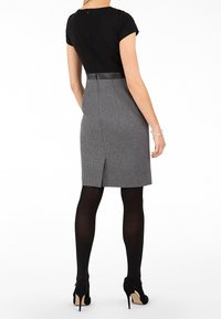 Robe en mélange de laine grise avec un haut noir ajusté, des manches courtes et une fermeture éclair sur le côté. Présente une fente discrète à l'arrière. Portée avec des collants noirs et des talons.