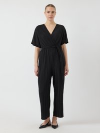 Žena oblečená v čiernom plisovanom zaviazanom overal s krátkymi rukávmi a čiernymi špicatými slingback balerínami, stojaca na bielom pozadí.