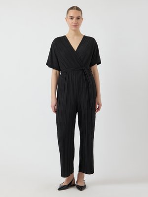 Kvinne iført en svart plisserte omslags-jumpsuit med korte ermer og svarte spisse slingback-sko, stående mot en hvit bakgrunn.