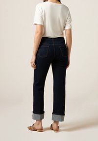 Woman wearing white short-sleeve top, dark cuffed jeans, and embellished tan slip-on shoes, standing against a plain light background.