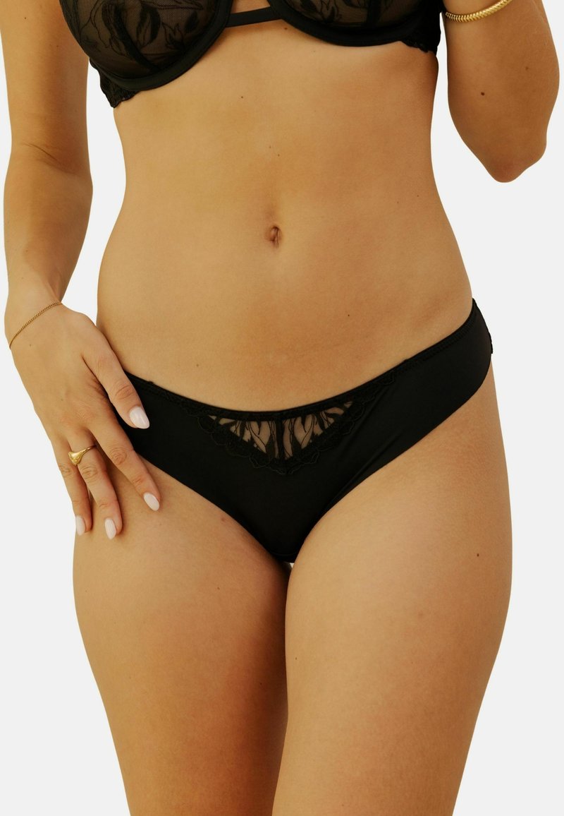 Bikini en dentelle noire avec un détail floral transparent à l'avant. Tissu doux, coupe basse et texture lisse. Ajustement autour des hanches.