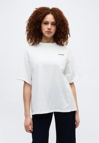 Νεαρή γυναίκα με σγουρά μαλλιά που φοράει μια φαρδιά λευκή μπλούζα Levi’s και σκουρόχρωμο παντελόνι, στέκεται μπροστά σε ένα απλό φωτεινό φόντο.