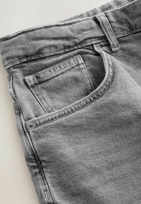 Primo piano di jeans in denim grigio che mostra la tasca anteriore, la piccola tasca porta monete, il passante per cintura e i dettagli delle cuciture.