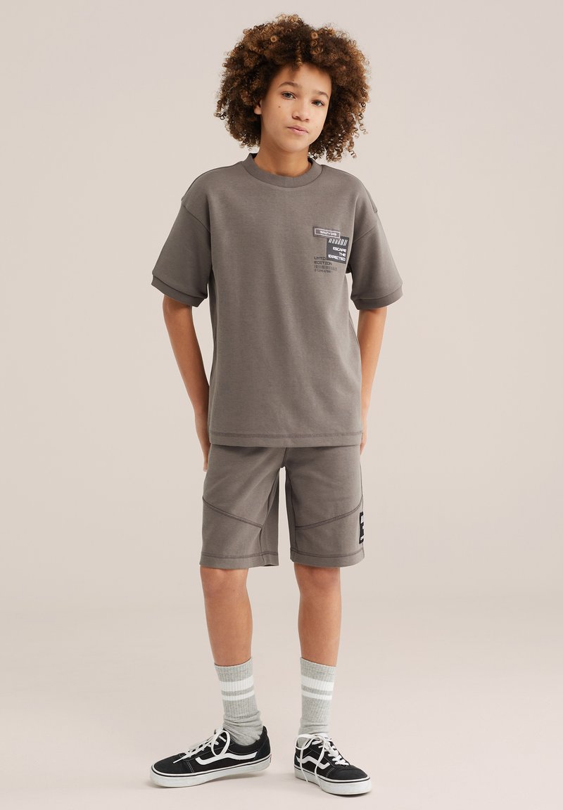 Băiat cu păr creț purtând un tricou gri larg și pantaloni scurți, șosete gri cu dungi și adidași negri, stând în picioare pe fundal simplu.