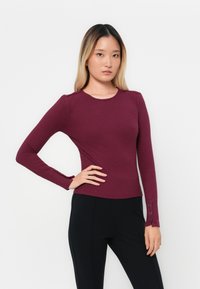 Haut long à manches ribbed bourgogne avec un col rond et des détails de boutons sur les manches, assorti à un pantalon noir. Texture lisse.