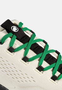 Κοντινή λήψη λευκού αθλητικού παπουτσιού με μαύρο γλωσσίδι και πράσινα κορδόνια που περνούν μέσα από μαύρους κρίκους σε υφασμάτινο φάκελο με υφή.