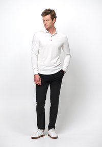 Weißes langärmliges Polohemd mit Kragen, drei schwarzen Knöpfen, kombiniert mit schwarzen eng anliegenden Hosen und weißen Sneakers vor einem neutralen Hintergrund.