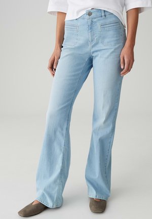 Persona che indossa jeans a zampa d'elefante azzurri chiari, top bianco e scarpe basse marroni, in piedi contro uno sfondo grigio semplice.