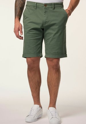 Mand iført grønne shorts med opbukket kant og hvide sneakers, stående med den ene hånd i lommen mod en ensfarvet baggrund.