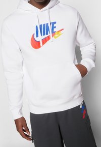 Vit huvtröja med en flerfärgad Nike-logotyp (röd, blå, gul) på bröstet, med en känguruficka och ribbade muddar.