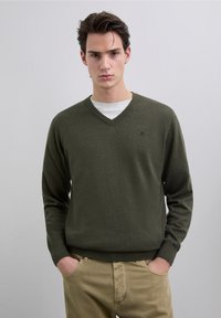 Suéter verde oliva con cuello en v, hecho de material suave, con mangas largas y un corte ajustado. Pequeño logo en el pecho, combinado con pantalones beige.
