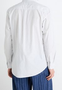 Camicia leggera con bottoni, caratterizzata da righe verticali bianche e grigio chiaro, con orlo curvo e polsini abbottonati. Tessuto dalla consistenza liscia.