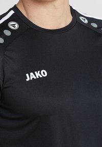 Černé sportovní triko vyrobené z materiálu odvádějícího vlhkost. Na přední straně se nachází bílé logo "JAKO" a pruhované akcenty na ramenech.