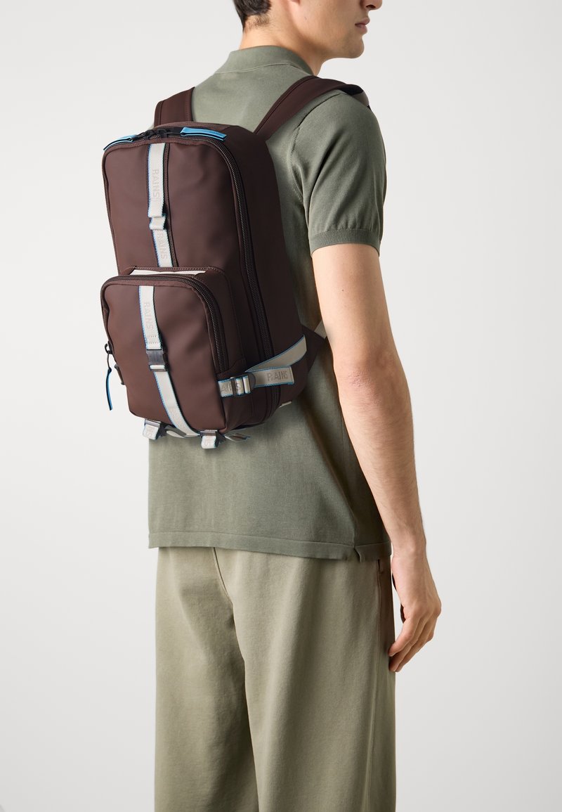 Homme portant un polo vert olive et un pantalon avec un sac à dos marron et gris comportant plusieurs compartiments zippés et des sangles.