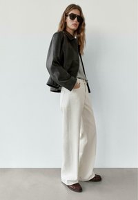 Svart lädercroppad jacka över en grå topp, vita vida byxor, mörkbruna loafers och stora svarta solglasögon. Mjuka texturer.