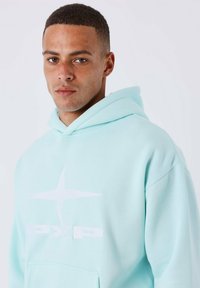 Sweatshirt à capuche vert menthe fabriqué en tissu doux. Il présente un logo blanc et dispose d'une poche kangourou. Design simple avec une coupe décontractée.