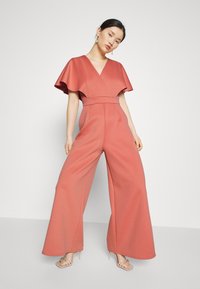 Κοραλί ολόσωμη φόρμα με μακριά πόδια και V λαιμόκοψη, με φτερωτές μανίκες, φτιαγμένη από λείο ύφασμα, featuring σφικτή μέση και πλαϊνές τσέπες.