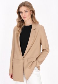 Beige puku muotoilulla, jossa on yksi nappi, kaarevat kaulukset ja kaksi etutaskua. Kiinnitetty mustan paidan päälle.
