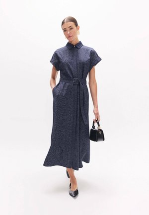 Femme portant une robe midi bleu foncé ceinturée avec un imprimé animal discret, tenant un sac à main noir et portant des chaussures noires pointues.