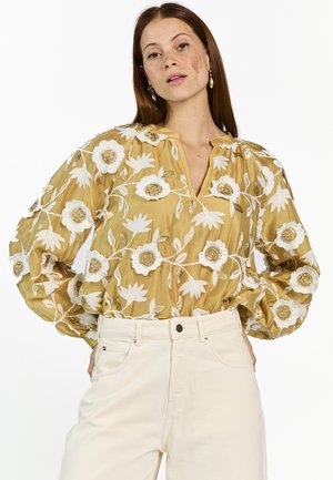 Frau mit langen roten Haaren, die eine goldene Bluse mit weißer Blumenstickerei und hoch taillierte, elfenbeinfarbene Hosen vor einem weißen Hintergrund trägt.