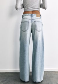 Personne portant un jean large bleu clair et un top court gris à manches longues, debout dos à la caméra contre un mur blanc.