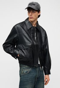 Νέος άνδρας που φοράει μαύρο δερμάτινο μπουφάν, μπλε τζιν και γκρι καπέλο, στέκεται με το ένα χέρι στην τσέπη μπροστά σε ουδέτερο φόντο.