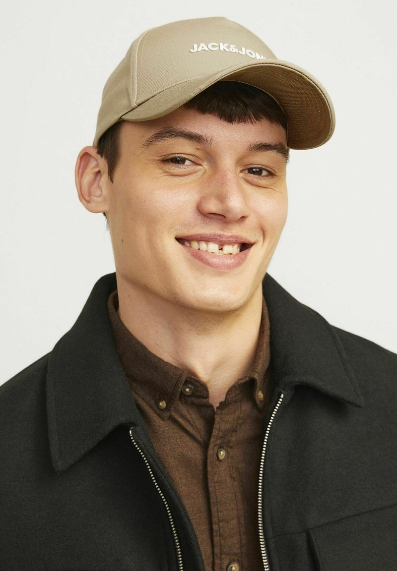 Beżowa czapka z logo "JACK&JONES", noszona przez osobę w ciemnej kurtce na brązowej koszuli zapinanej na guziki, z wyraźnymi rysami twarzy.