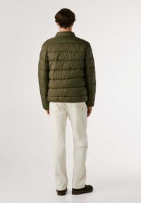 Doudoune vert olive avec des coutures horizontales, col mandarin et deux poches latérales, portée avec un pantalon blanc et des chaussures foncées.