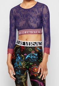 Top corto in rete blu navale con maniche lunghe in pizzo e marchio "Versace Jeans Couture". Abbinato a leggings multicolori con motivi floreali.