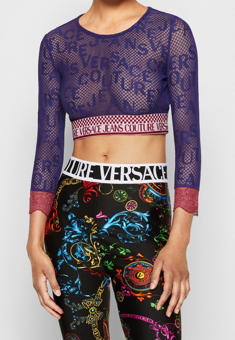 Top corto in rete blu navale con maniche lunghe in pizzo e marchio "Versace Jeans Couture". Abbinato a leggings multicolori con motivi floreali.