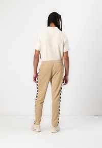 Isik, kellel on pikad palmikud, kannab valget lühikeste varrukatega särk, beeže pükse tumesinise ja valge küljeribaga ning beeže tossusid, näoga ära.