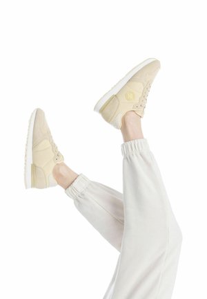 Zapatillas deportivas beige con detalles de ante y malla, suela blanca y logotipo en el lateral, combinadas con pantalones de chándal claros, con los pies elevados.