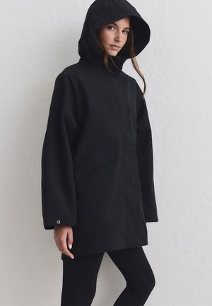 Femme portant une veste à capuche noire et un pantalon noir, debout contre un mur blanc texturé, regardant vers la caméra.