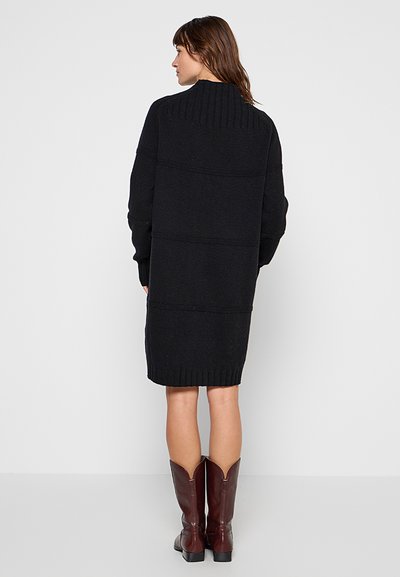 Femme vue de dos portant une longue robe pull noire et des bottes en cuir marron foncé mi-mollet, sur un fond uni.