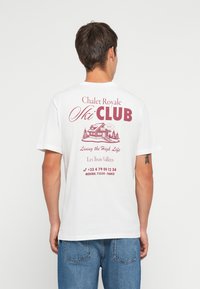Hvit bomull t-skjorte med et maroon grafisk design på baksiden, inkludert en hytte, tekst og et telefonnummer for en skiklubb.