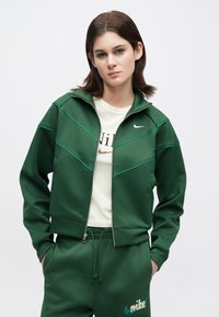 Νέα γυναίκα που φοράει σκούρο πράσινο φούτερ με φερμουάρ Nike και αντίστοιχα φόρμες, στέκεται με τα χέρια στις τσέπες σε έναν απλό λευκό φόντο.