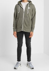 Grauer Zip-Hoodie mit weißen Kordeln und Logo, kombiniert mit dunklen Jeans und grauen Sneakers. Einfaches Design, weiche Textur und sportlicher Schnitt.