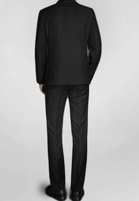 Costume noir avec une coupe ajustée, tissu lisse, pantalon droit, fente simple à l'arrière, associé à des chaussures noires.