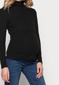 Svart ribbad graviditetsturtleneck med en åtsittande bodice, design ovanför magen, och långa ärmar, i kombination med blå jeans.
