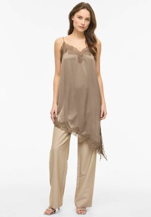 Jeune femme portant une robe nuisette en satin taupe avec bordure en dentelle sur un pantalon beige à jambes larges et des talons ouverts argentés, debout devant un fond blanc.