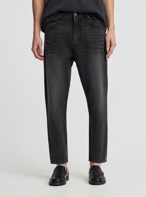 Persona che indossa jeans neri slavati a gamba dritta e scarpe slip-on nere, in piedi contro uno sfondo chiaro e uniforme.