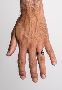 Anello in metallo nero con un design moderno, che mostra la forma di un'ancora. La mano ha una tonalità di pelle abbronzata e vene visibili, con unghie corte e di colore chiaro.