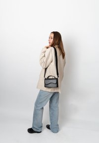 Femme en blazer beige et jean clair se tenant de profil, portant un sac bandoulière noir avec une large bandoulière sur un fond blanc uni.