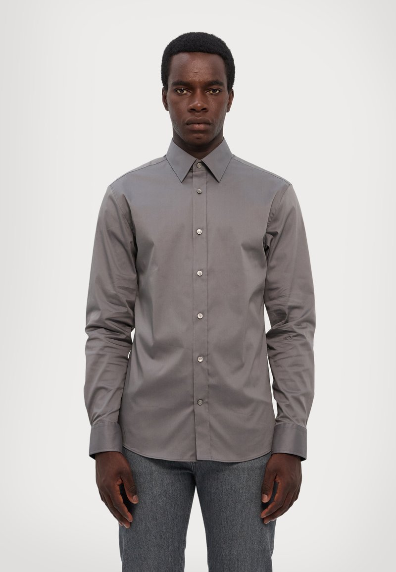 Chemise grise à col classique et à manches longues. Présente une texture lisse et une coupe ajustée, avec des boutons visibles sur le devant.