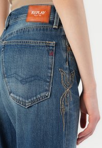 Denim jeans met een donkerblauwe wassing. Heeft borduring op de achterzak en een bruine leren label op de taille, wat de merkidentiteit benadrukt.